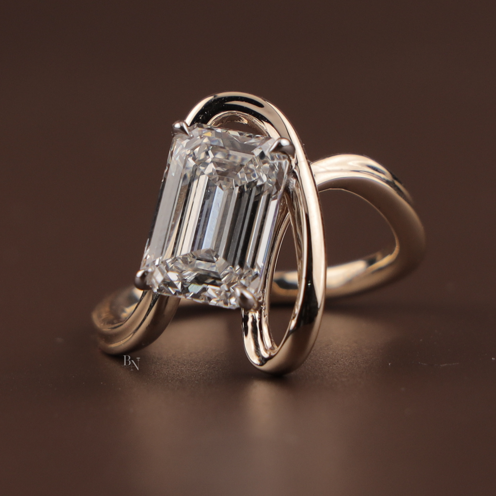 4 carat emerald cut lab grown diamond set horizontally in a modern east-west tension swirl bypass setting of polished 14k rose gold, shown on a hand to highlight the large step-cut facets and unique architectural design.