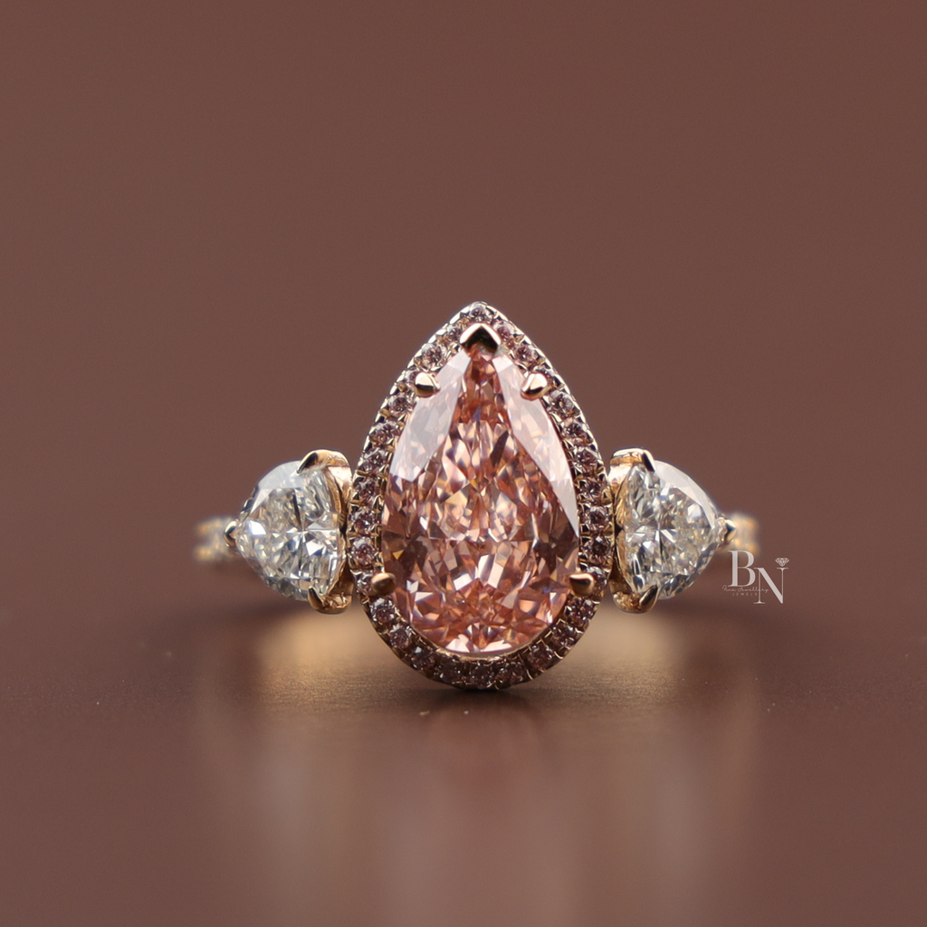 Close up view of a 2 carat teardrop pear cut fancy pink lab grown diamond engagement ring in 14k rose gold surrounded by a micro pave halo framework flanked by clear pear side accent gems on a diamond lined band.