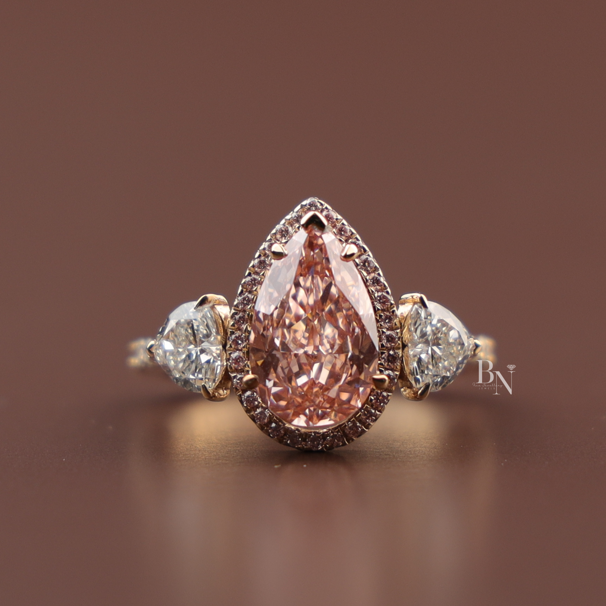 Close up view of a 2 carat teardrop pear cut fancy pink lab grown diamond engagement ring in 14k rose gold surrounded by a micro pave halo framework flanked by clear pear side accent gems on a diamond lined band.