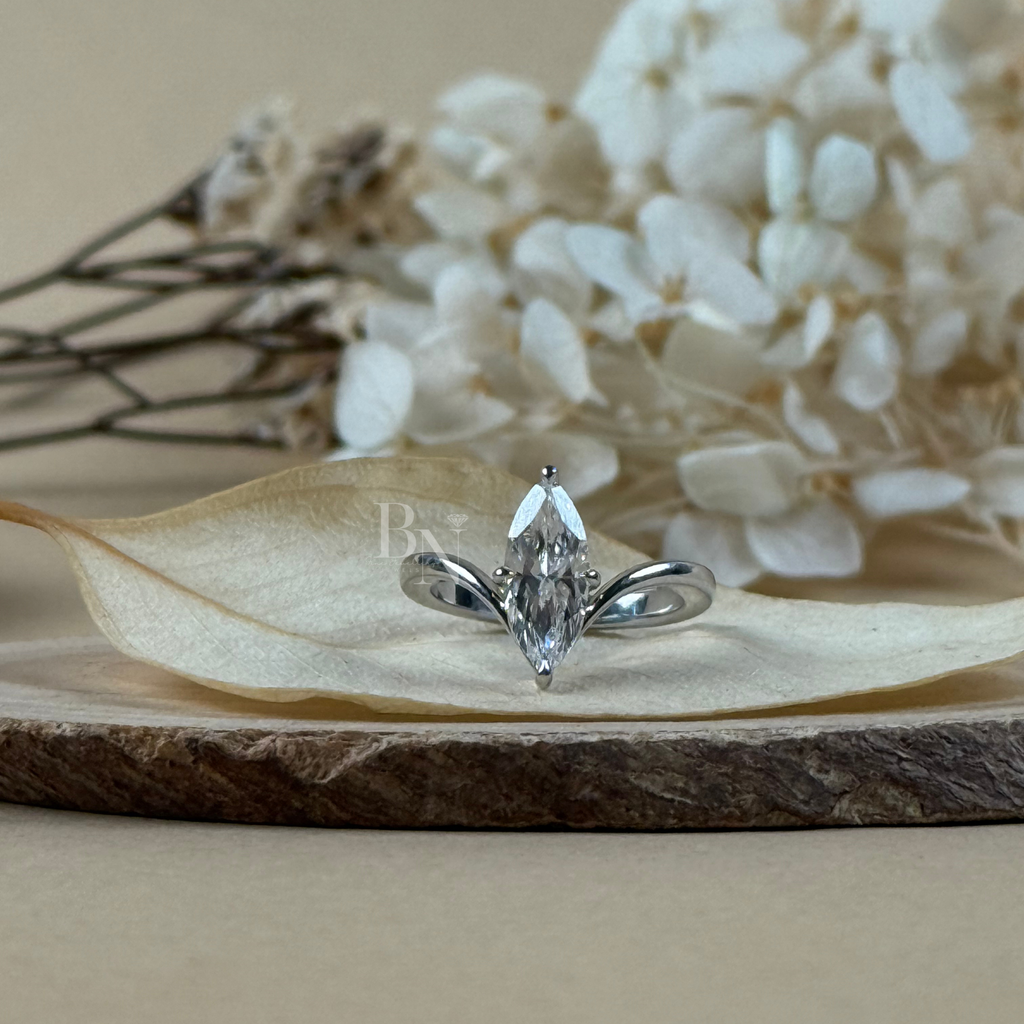 May include: A marquise-cut diamond engagement ring with a silver band. The diamond is set in a prong setting. The ring is displayed on a wooden surface with dried flowers in the background. The ring is a symbol of commitment.