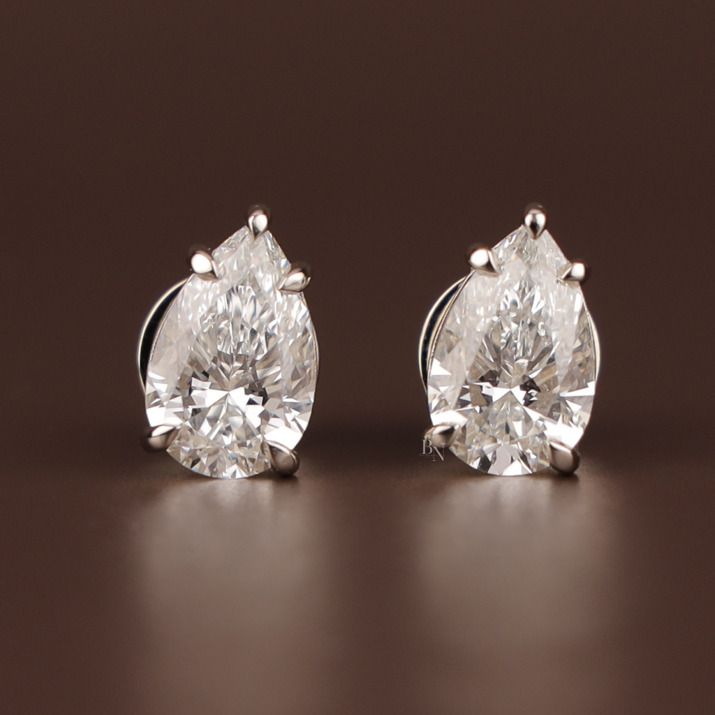 May include: A pair of pear-cut diamond stud earrings. Each earring showcases a clear, faceted diamond set in a silver-coloured metal. The diamonds are the focal point, reflecting light against a dark brown background.