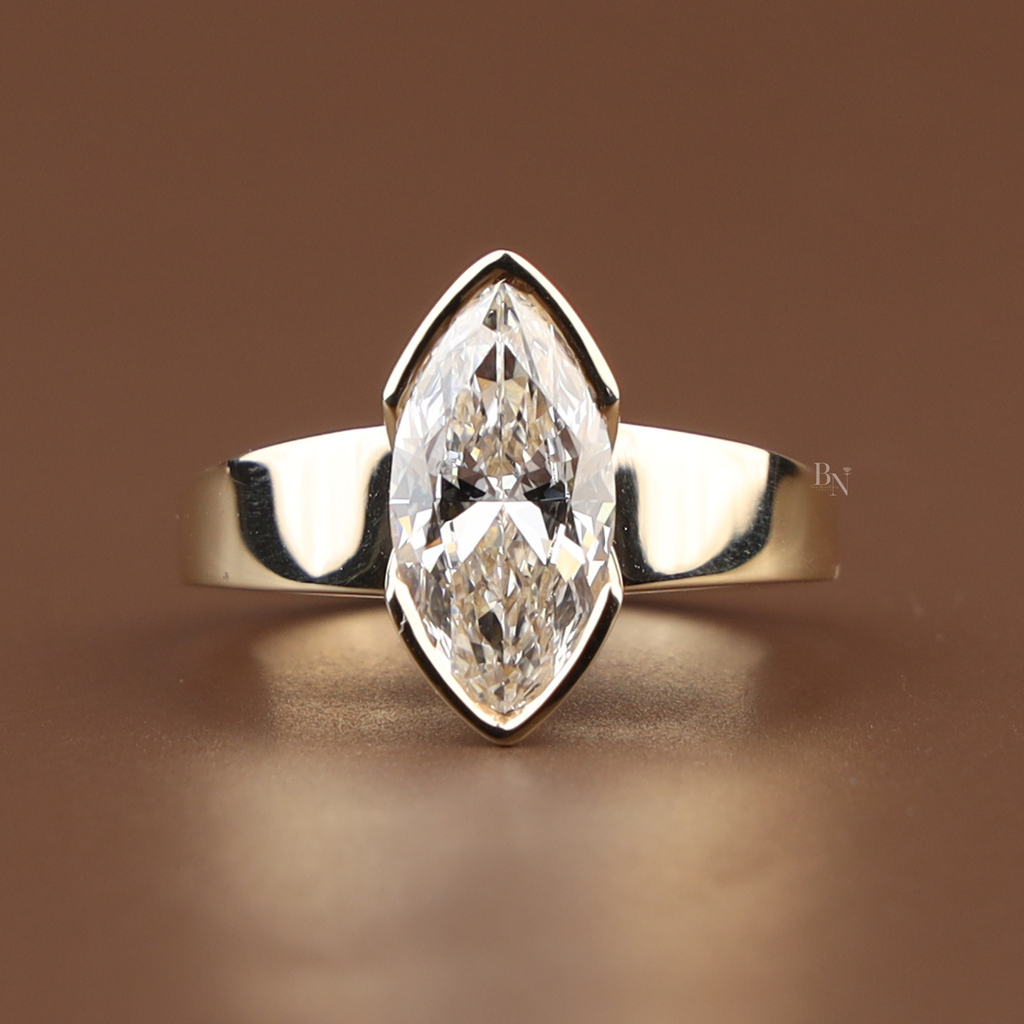 Hand shot of a 2 carat marquise cut lab grown diamond engagement ring in 18k yellow gold featuring a modern half-bezel setting with a thick tapered flat band and architectural V-shaped tips.