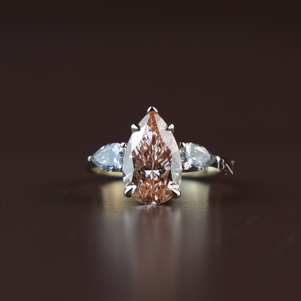 Three stone engagement ring featuring a 3 carat fancy pink pear cut lab grown diamond center stone with protective claw prongs, flanked by two white pear cut side diamonds on a tapered 14k yellow gold band.