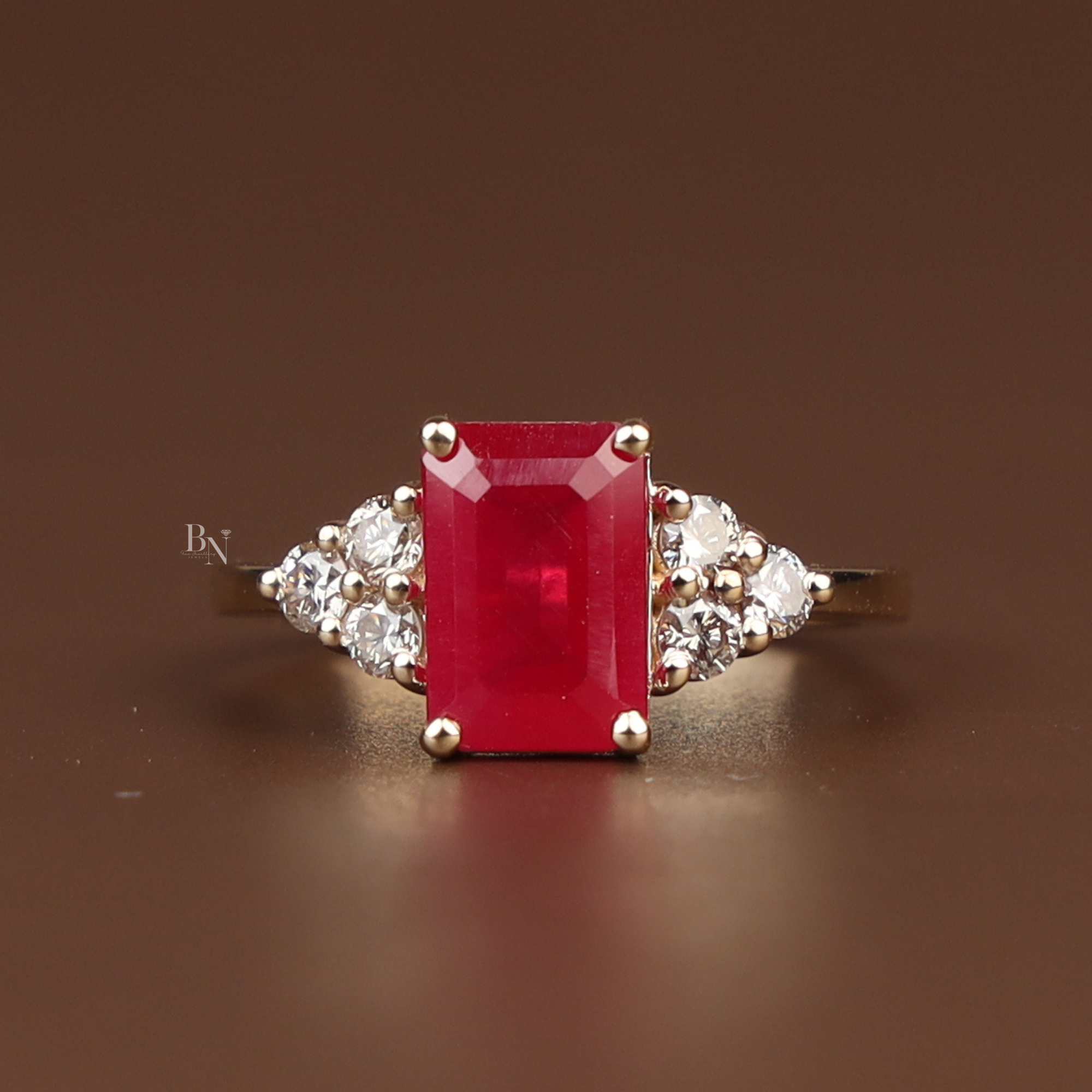 Close up of 2 carat emerald cut red lab diamond engagement ring in 14k yellow gold featuring a hidden halo under the center stone and triple diamond cluster side accents on a polished gold band with an opaque pigeon blood red stone.