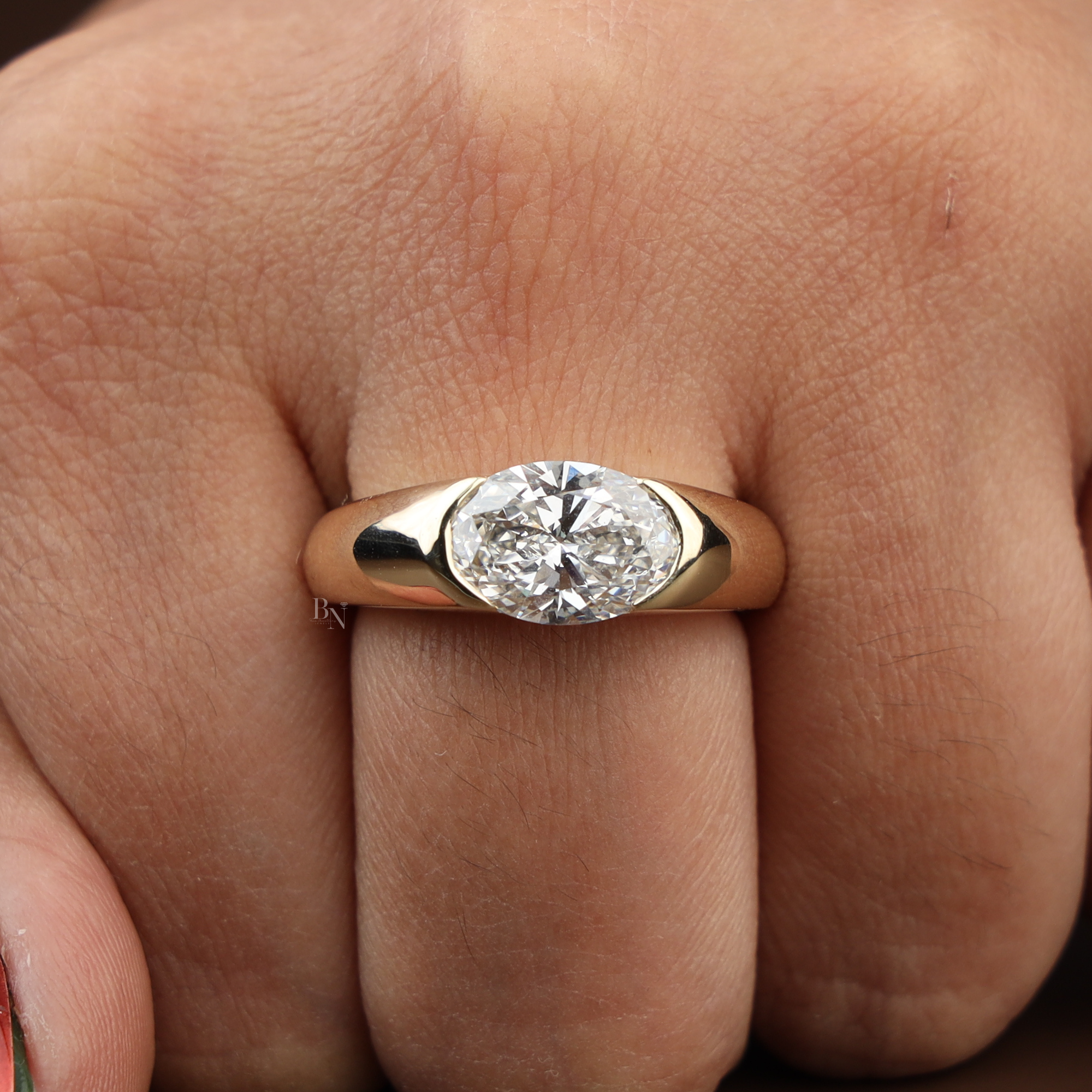 Macro view of a 2.50 carat oval lab grown diamond set horizontally in a 14k yellow gold half-bezel signet engagement ring with a thick polished tapered band.