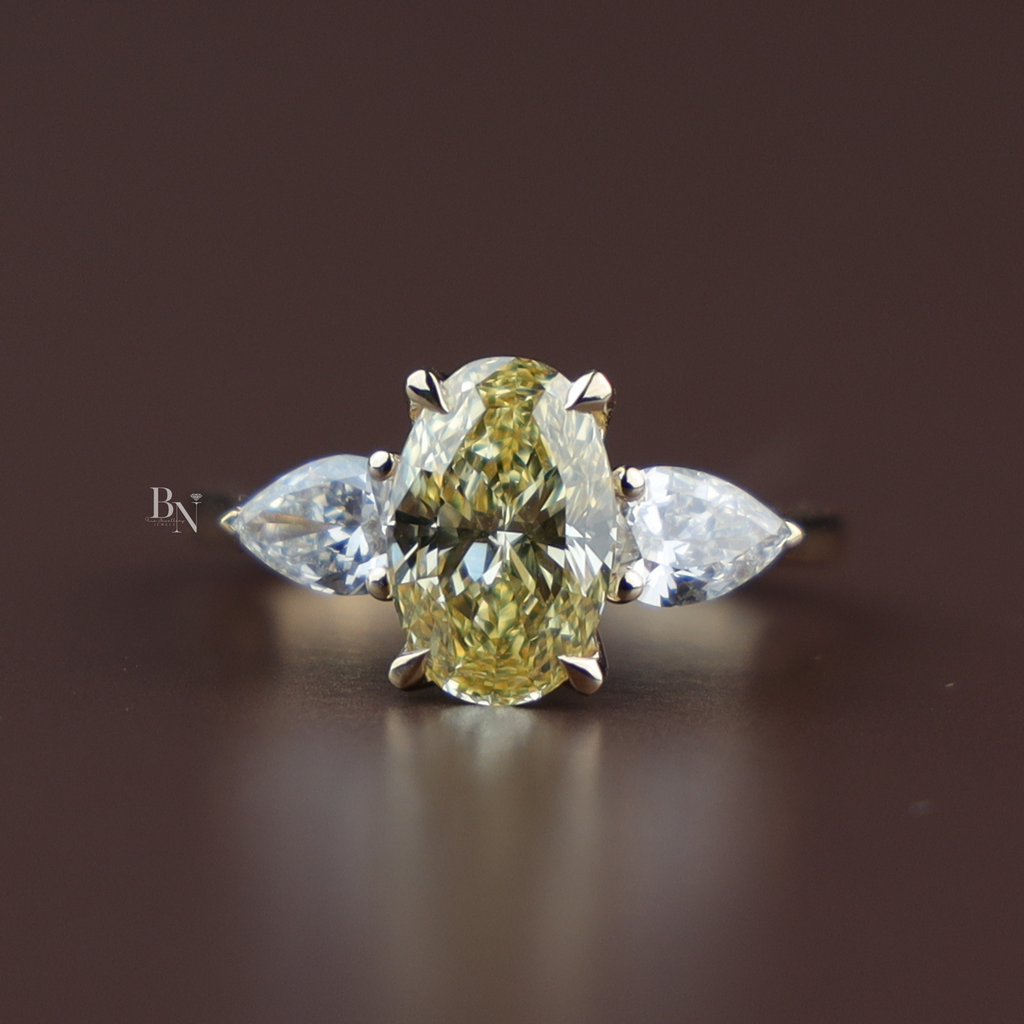 Close up view of a 2 carat elongated oval cut fancy canary yellow lab grown diamond engagement ring in 14k yellow gold flanked by two clear pear cut side stones on a dainty polished cathedral band configuration.