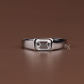 May include: A silver ring featuring a rectangular, clear gemstone. The gemstone is set in a bezel setting, and the band is thick and polished. The ring is displayed on a brown surface.