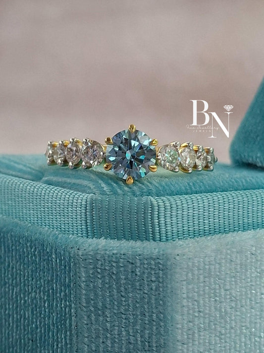A 1-carat fancy vivid blue diamond engagement ring shown in its velvet box. The blue diamond is set in a 14k yellow gold band with graduated white diamonds on the side