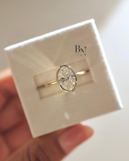 A modern 2 carat oval lab-grown diamond ring in a white ring box. This ethical solitaire ring features a sleek yellow gold bezel setting and a thin band, creating a chic, minimalist and durable look perfect for everyday wear.