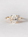 An artistic 2 carat oval lab-grown diamond engagement ring on a finger. This ethical, nature-inspired ring is crafted in 14K yellow gold and features a unique side cluster of pear and round lab diamonds for a boho look.