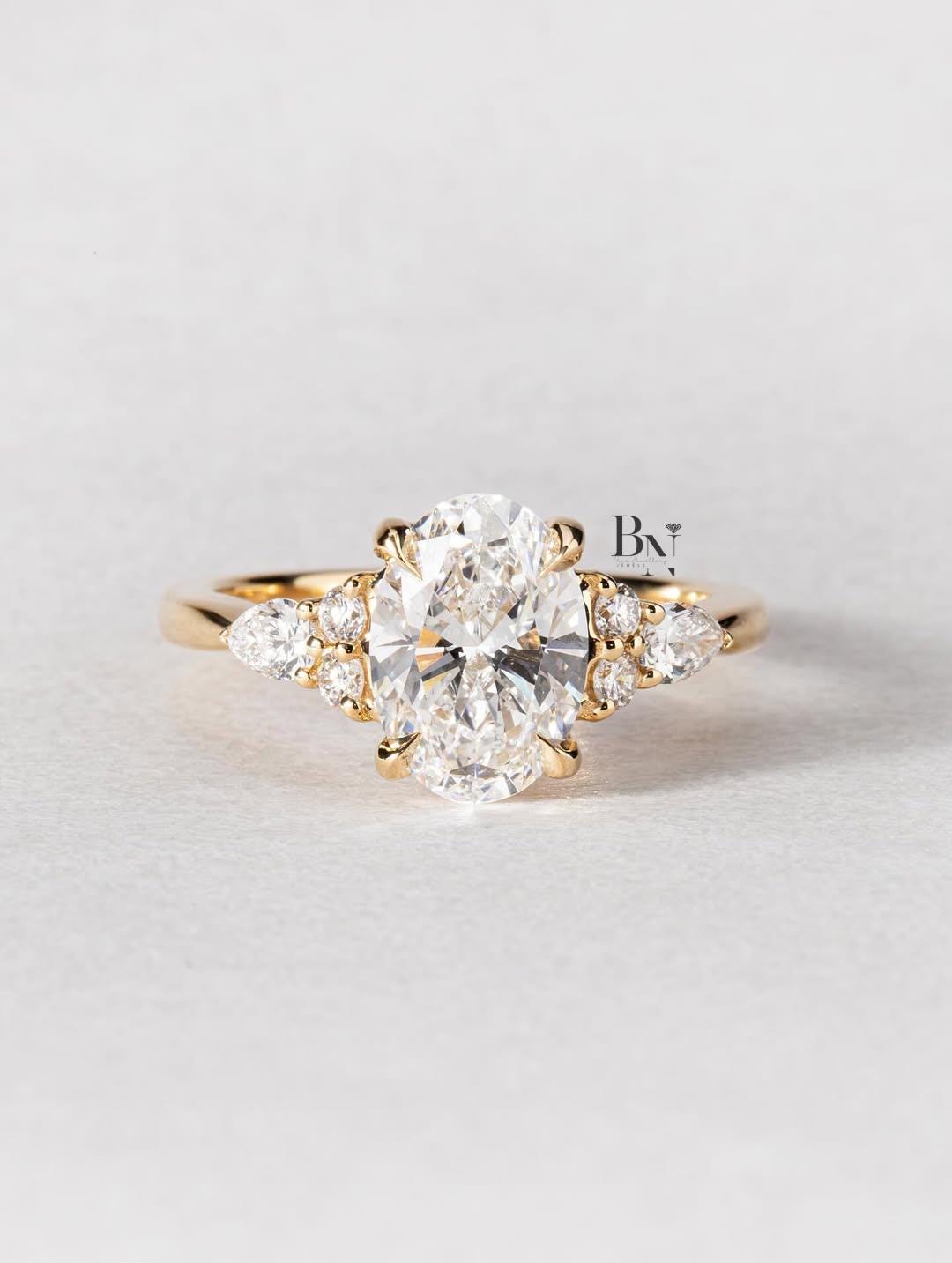 An artistic 2 carat oval lab-grown diamond engagement ring on a finger. This ethical, nature-inspired ring is crafted in 14K yellow gold and features a unique side cluster of pear and round lab diamonds for a boho look.