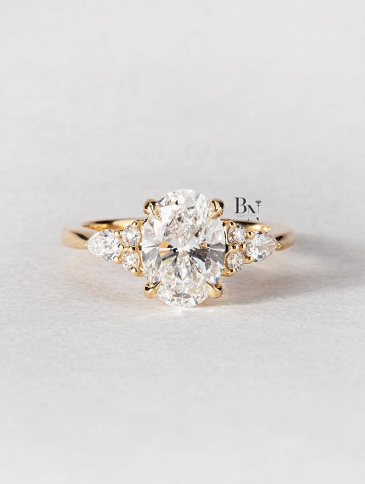 An artistic 2 carat oval lab-grown diamond engagement ring on a finger. This ethical, nature-inspired ring is crafted in 14K yellow gold and features a unique side cluster of pear and round lab diamonds for a boho look.