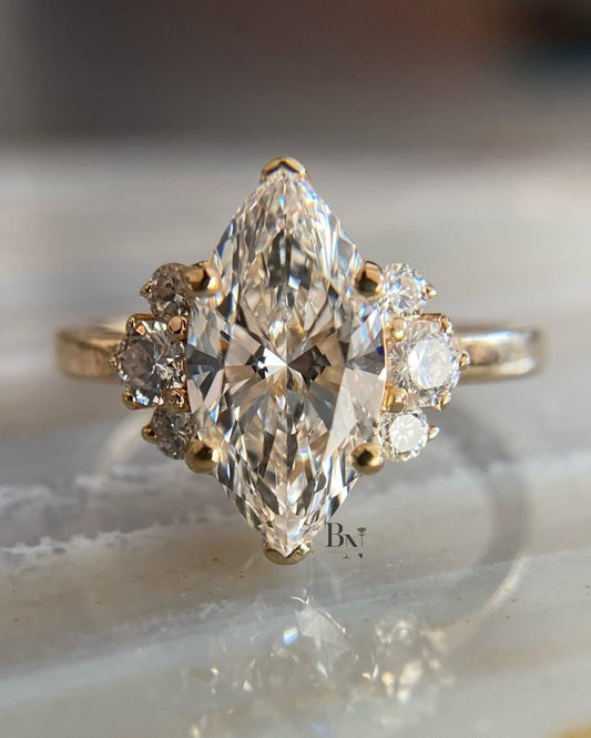 An artistic 2 carat marquise lab-grown diamond engagement ring on a finger. This ethical, nature-inspired ring is crafted in 14K yellow gold and features a unique side cluster of round lab diamonds for a boho look.