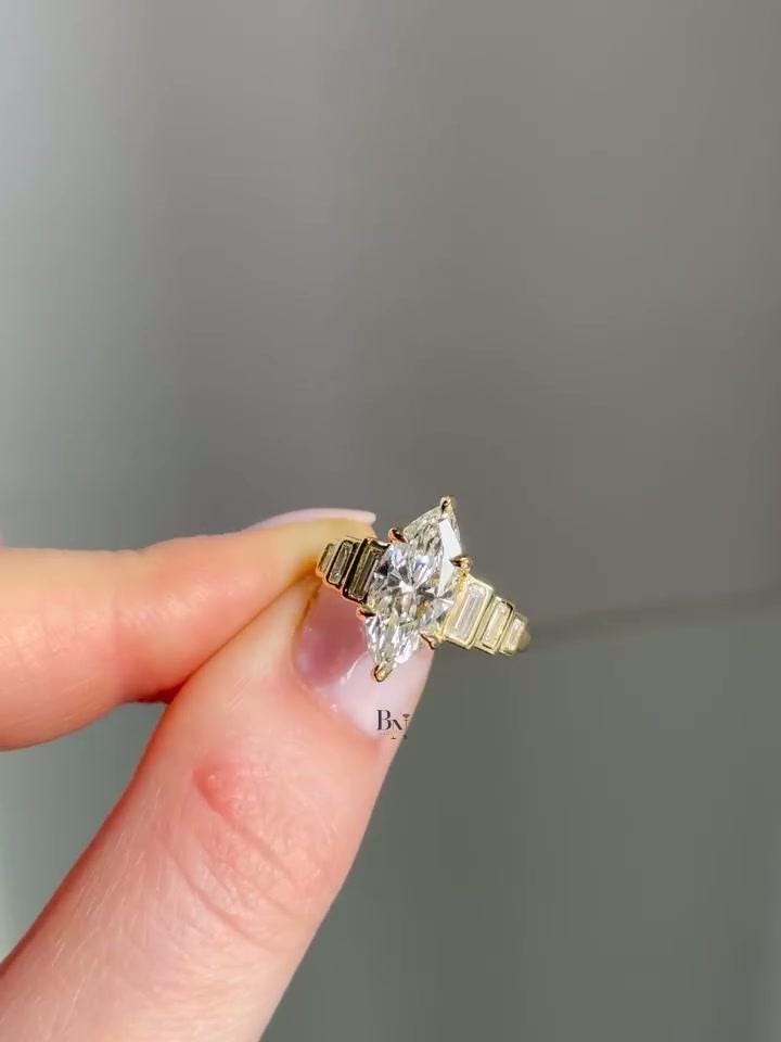A woman's hand wearing a 2 carat Art Deco marquise moissanite ring. The vintage-style ring features a brilliant marquise center stone flanked by graduated, channel-set baguette diamonds on a substantial 14K yellow gold band.