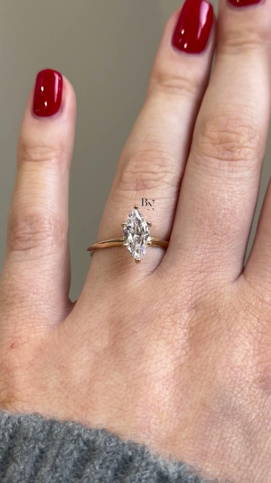 A classic 1 carat marquise lab diamond solitaire engagement ring against a neutral background. The IGI certified diamond is set on a delicate, thin yellow gold band, showcasing its minimalist and timeless design perfect for a promise or engagement.