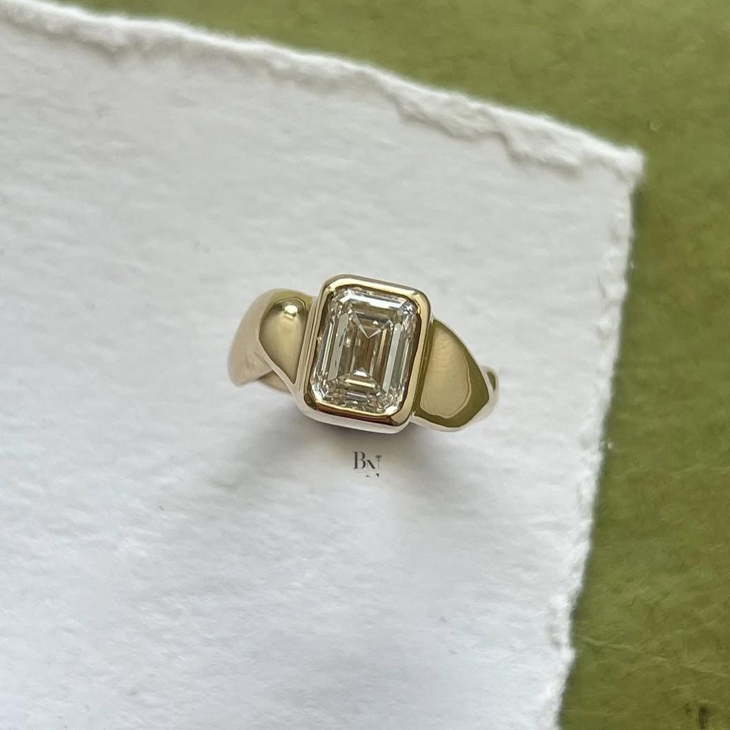 Close-up of 3 carat emerald cut lab grown diamond in a full bezel setting, showcasing the thick, modern yellow gold signet style band and clean step-cut clarity.